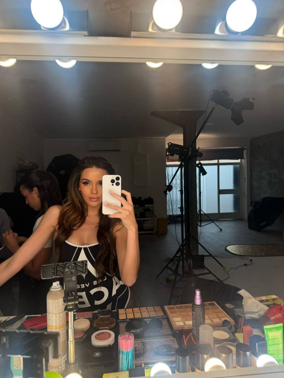 Professional beauty model taking a mirror selfie inside a luxury makeup studio, surrounded by premium cosmetics, makeup mirrors with Hollywood lights, backstage fashion photoshoot atmosphere, glamorous look, ultra realistic photography, 8K.