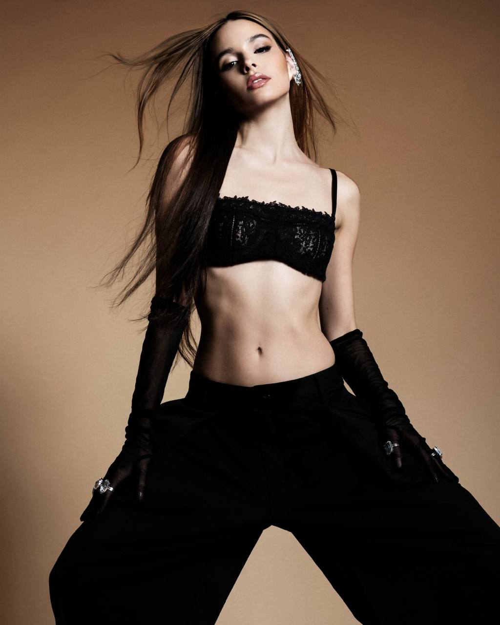 Ultra realistic full body portrait of a glamorous young woman with long silky brown hair wearing a black lace bandeau top, elegant long gloves and oversized black trousers, luxury fashion editorial, beige studio background, dramatic lighting, high fashion photography.