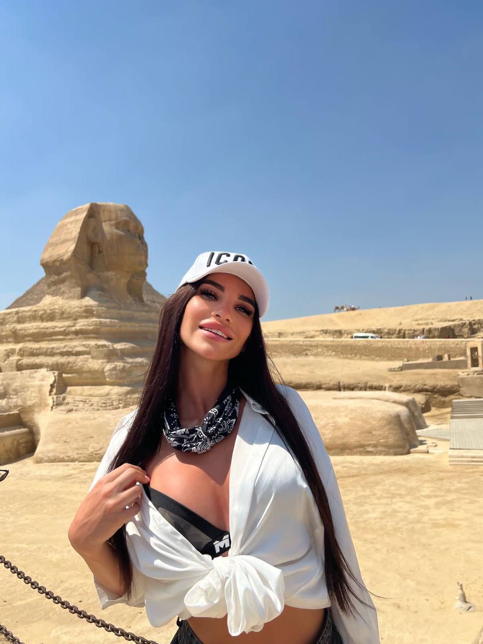 Smiling brunette in a white cap and knotted white shirt with a bandana posing in front of the Great Sphinx of Giza
