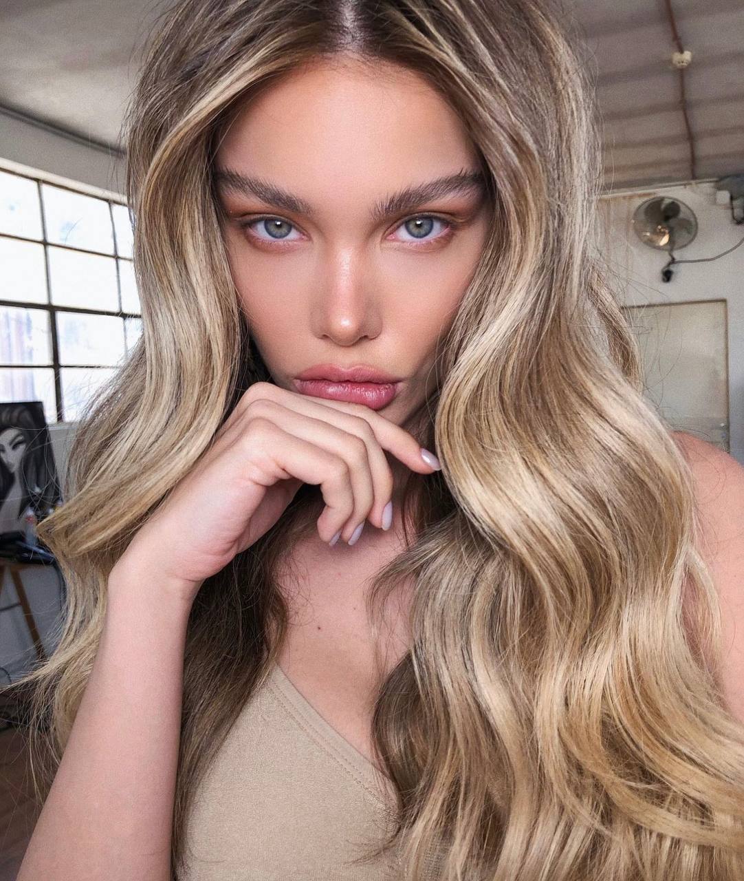 Close-up selfie of a woman with long wavy blonde hair, green eyes and natural makeup in a beige top