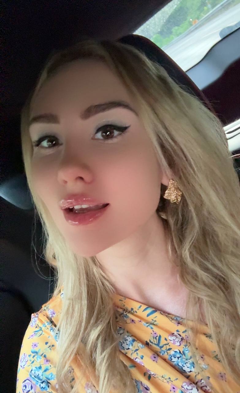 Ultra realistic close-up selfie of a glamorous blonde woman with long wavy platinum hair, flawless porcelain skin, glossy lips, winged eyeliner, wearing a yellow floral dress and elegant gold earrings, sitting inside a luxury car, soft natural lighting, lifestyle beauty portrait, RAW smartphone photo, ultra detailed, photorealistic, 8K.