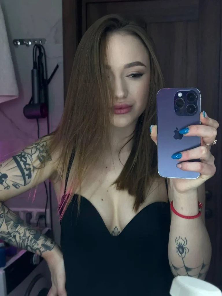 Mirror selfie of a woman with tattooed arms in a black top holding a purple phone