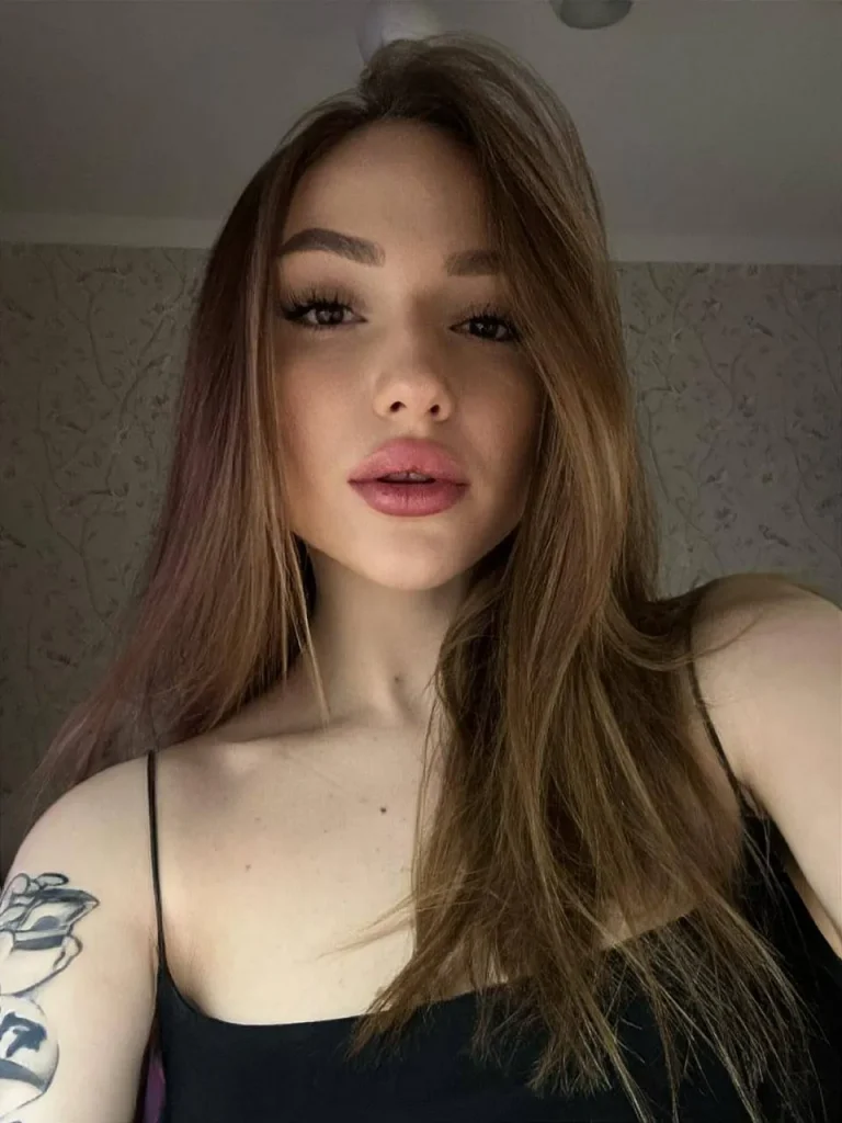 Close-up selfie of a young woman with long light brown hair and a shoulder tattoo in a black cami top