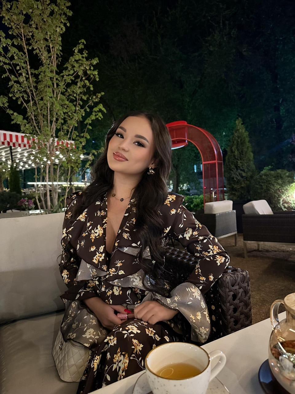 Ultra realistic seated portrait of a glamorous young woman with long silky jet black wavy hair, almond-shaped dark brown eyes, flawless glowing skin, wearing an elegant floral designer dress, sitting at a luxury outdoor café at night with a cup of tea, cozy ambient lighting, premium restaurant atmosphere, sophisticated lifestyle editorial, RAW DSLR photo, ultra detailed, photorealistic, 8K.