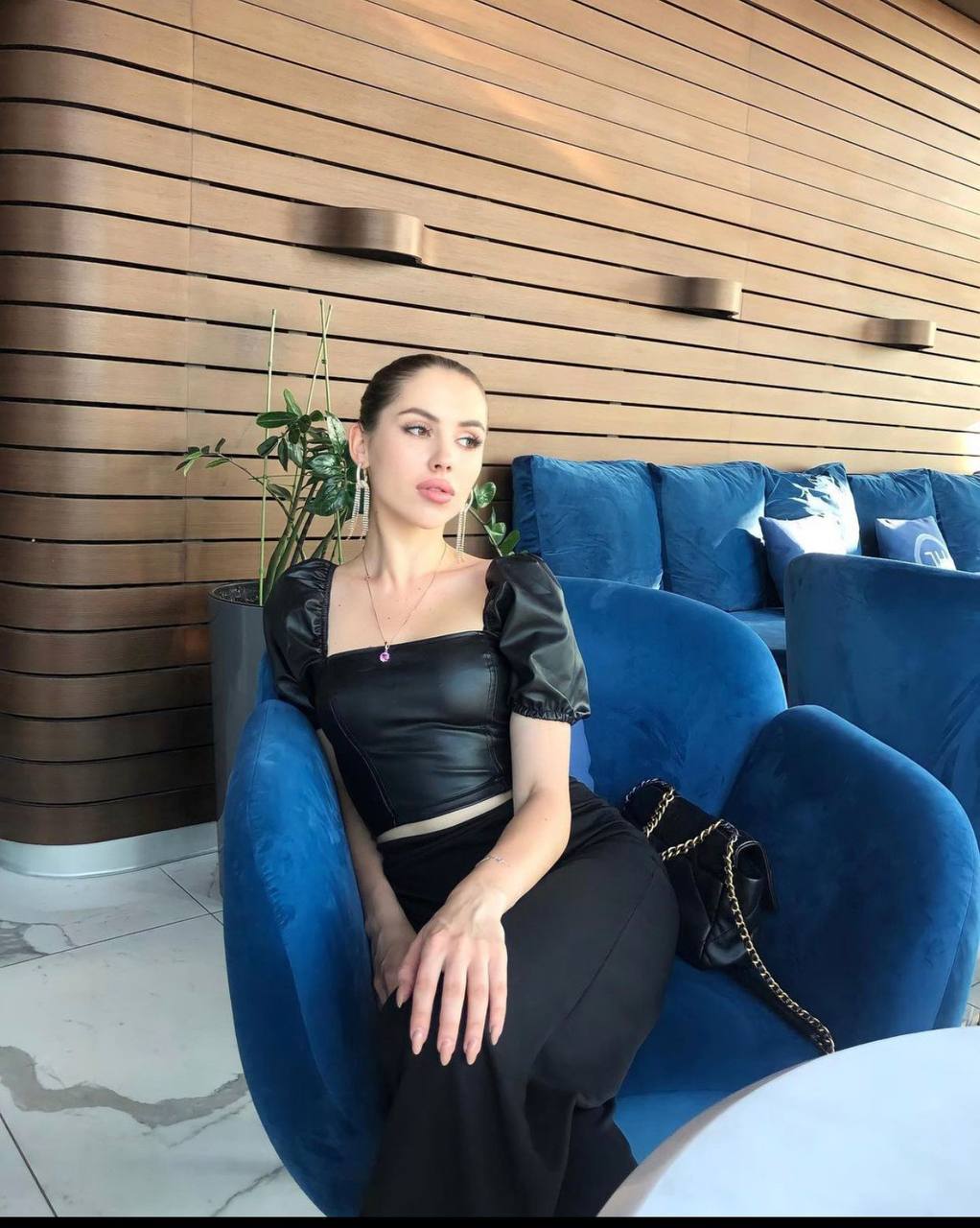 Woman with a sleek bun in a black faux leather puff-sleeve top and black trousers sitting in a blue velvet armchair