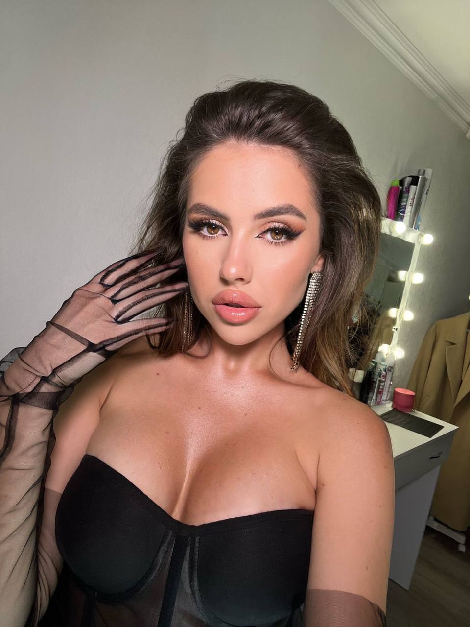 Close-up glam selfie of a brunette in a black strapless corset and sheer gloves with long crystal earrings
