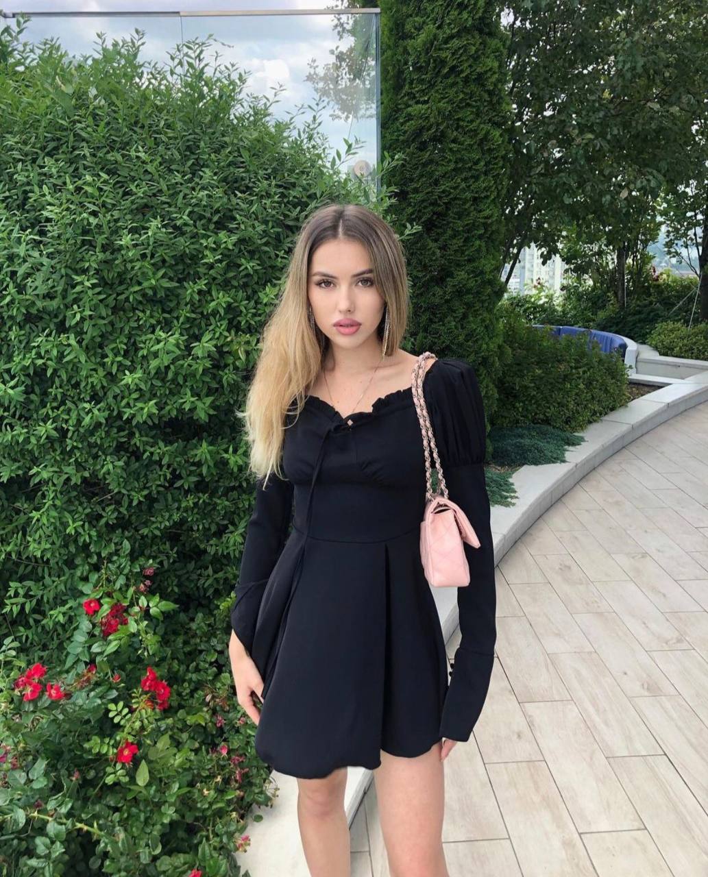 Blonde with ombre hair in a black puff-sleeve mini dress and a pink quilted shoulder bag standing on a garden path with red roses