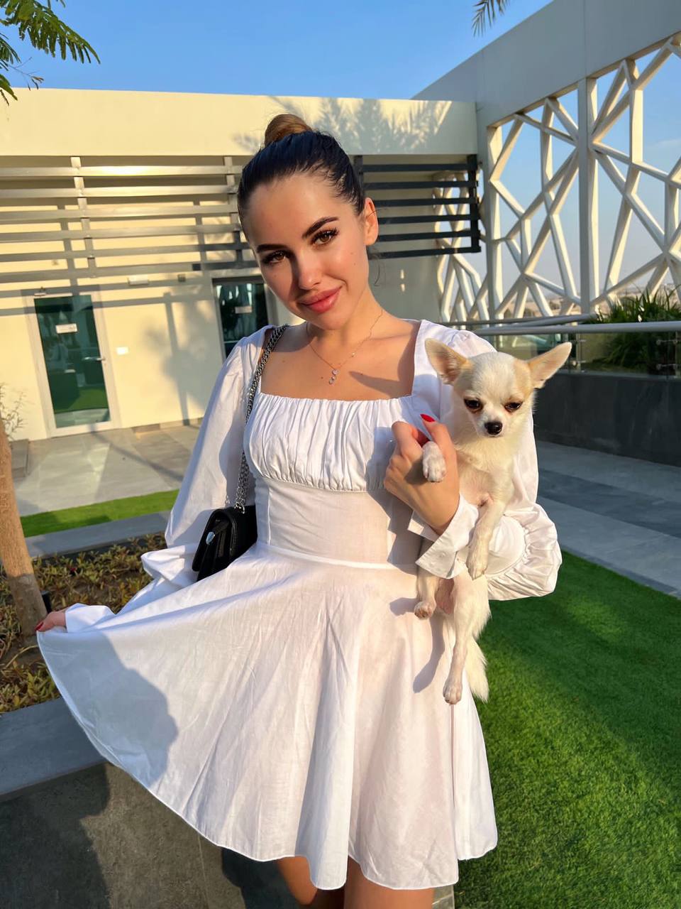 Smiling woman with a bun in a white puff-sleeve mini dress holding a white chihuahua outdoors at golden hour
