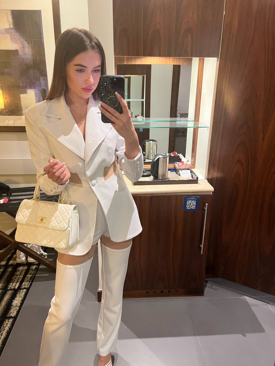Mirror selfie of a brunette in a white cut-out blazer suit with white thigh-high boots and a white quilted handbag