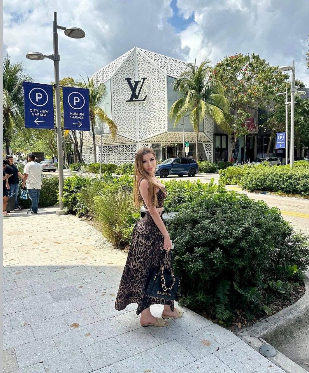 stylish brunette woman in leopard print maxi dress posing outside Louis Vuitton boutique with designer handbag