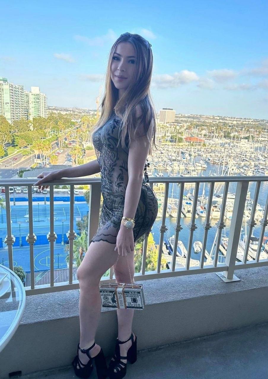 fashionable brunette woman in sheer black mini dress and high heels standing on hotel balcony overlooking marina