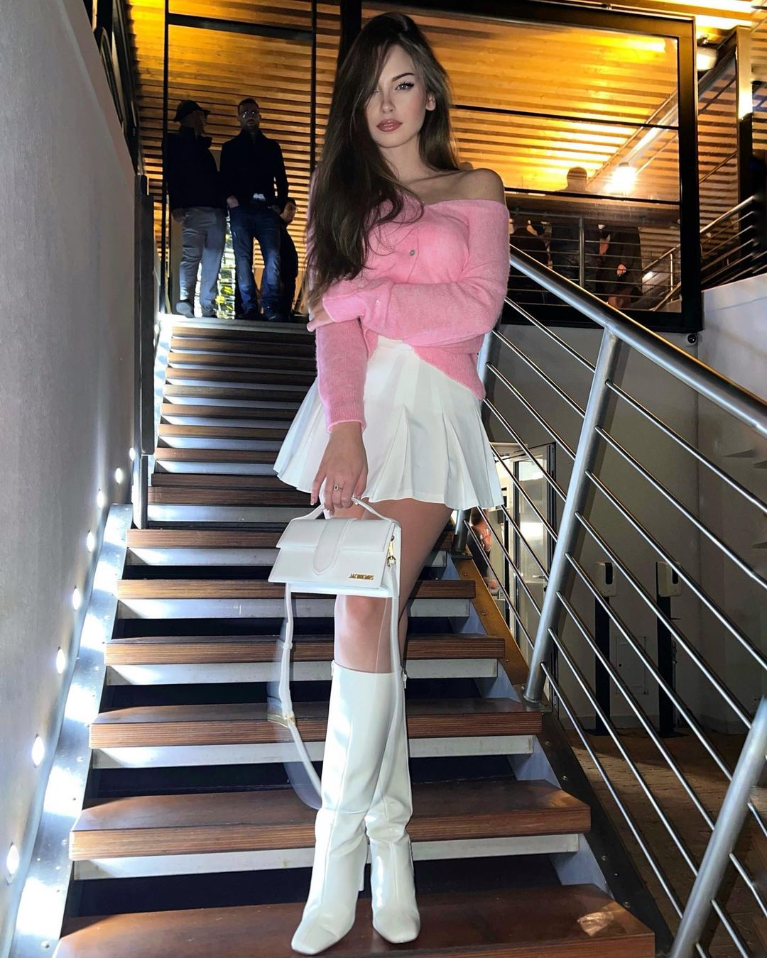 Brunette in a pink off-shoulder sweater, white pleated mini skirt and white knee-high boots posing on a staircase with a white handbag