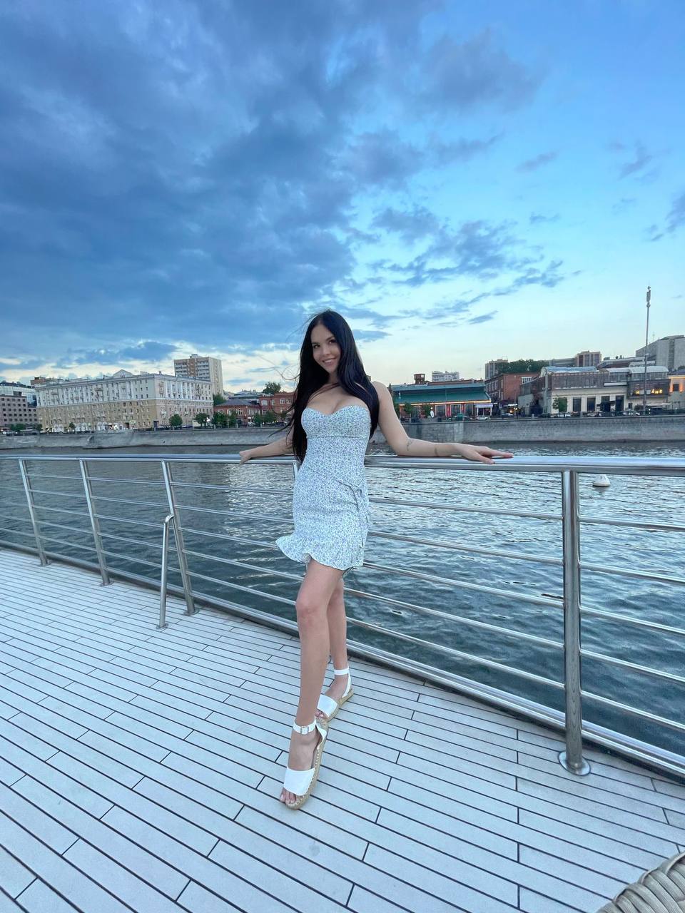 Brunette in short white floral summer dress and sandals posing by the railing on a river pier at dusk