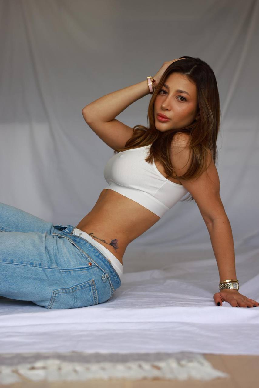 Beautiful brunette fashion model relaxing on white linen sheets wearing a white crop top and light blue jeans, cozy editorial aesthetic, natural daylight, luxury lifestyle campaign