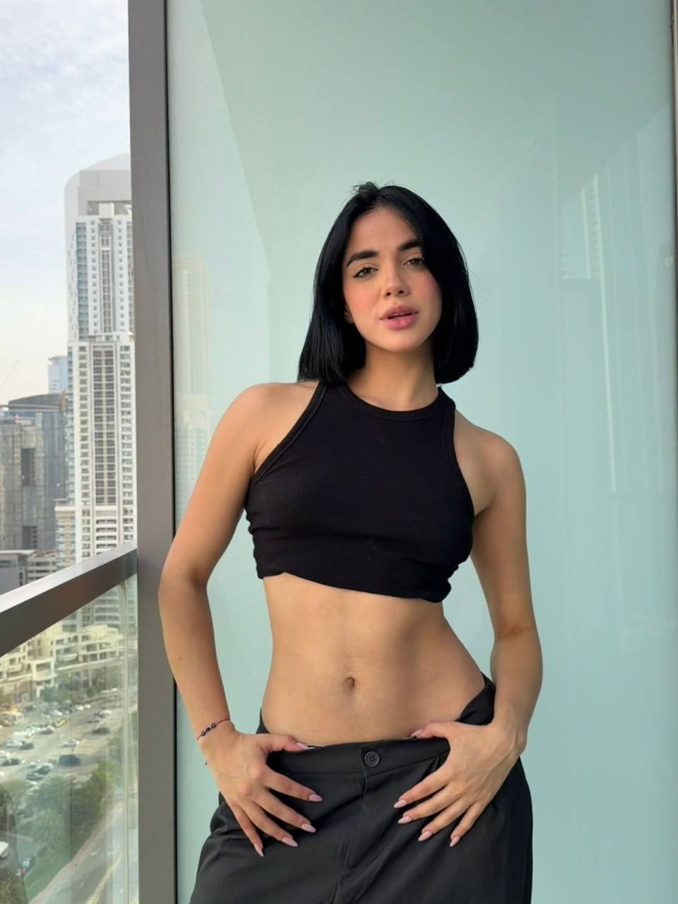 Luxury brunette escort model posing on a Dubai balcony wearing a black crop top with city skyline views