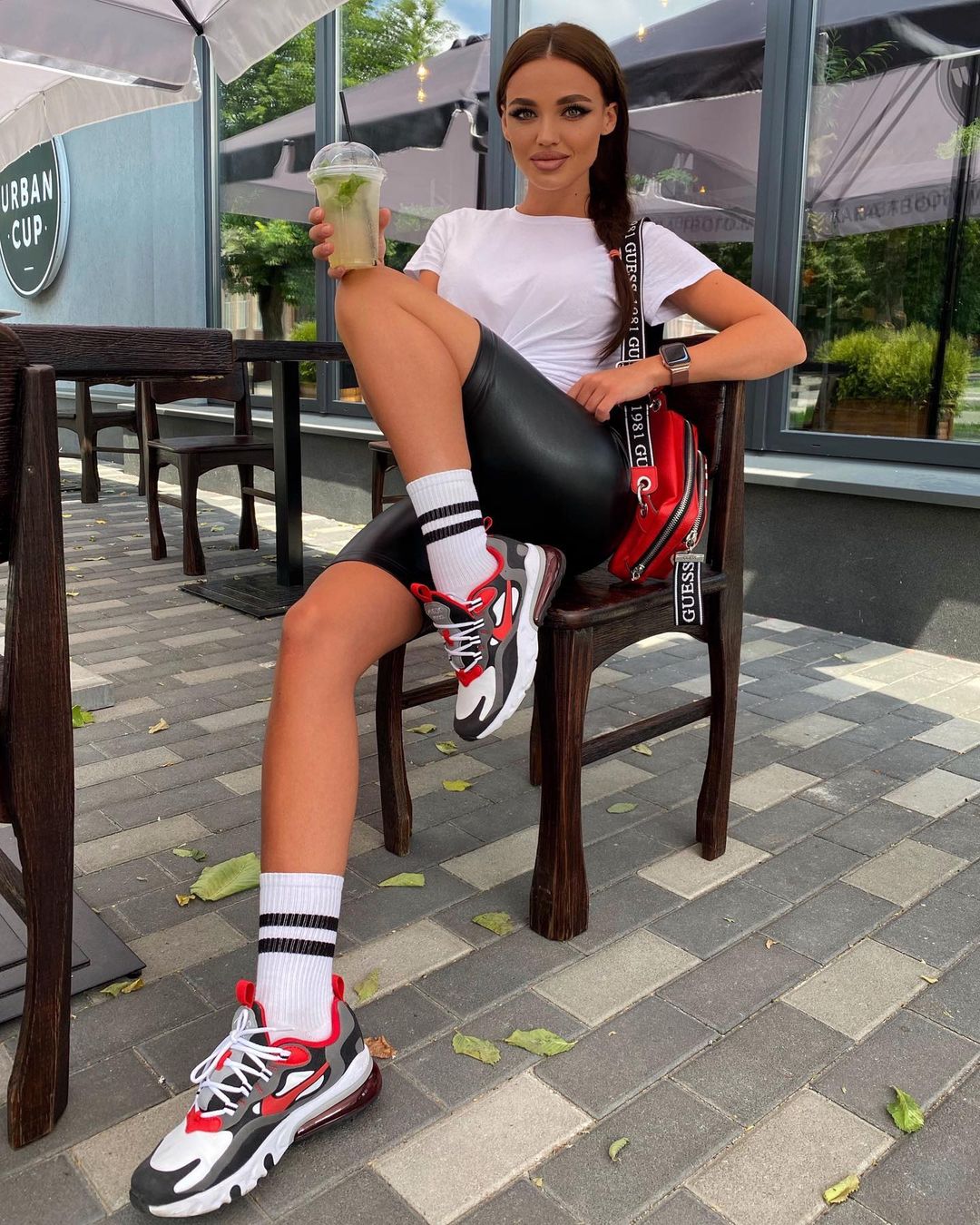 Brunette in a white t-shirt, black leather shorts and sneakers holding an iced drink at an outdoor cafe table with a red bag