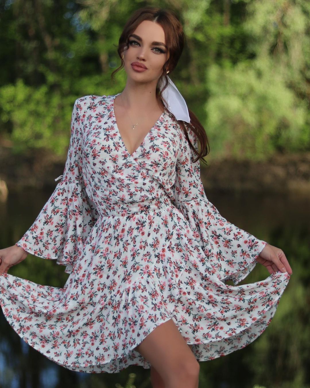 Woman with auburn hair and a white hair bow in a floral wrap mini dress with bell sleeves posing against green foliage