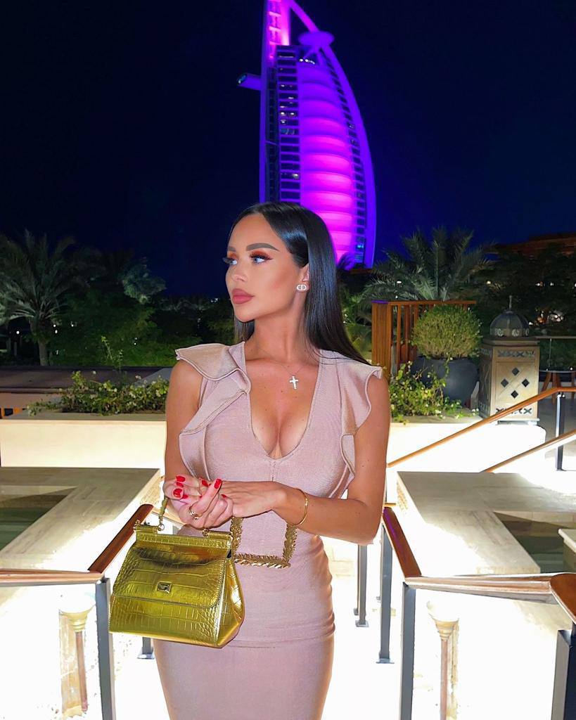 Brunette woman in a blush bodycon dress holding a gold handbag at night with the illuminated Burj Al Arab in the background