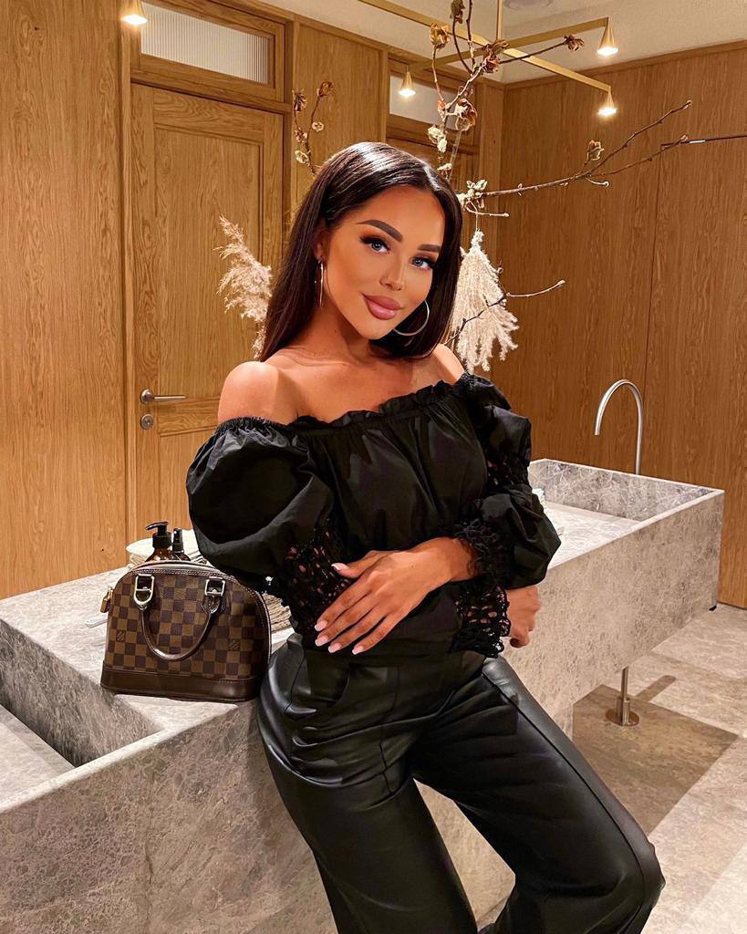 Woman in a black off-shoulder puff-sleeve top and leather trousers leaning on a marble counter with a brown checkered handbag