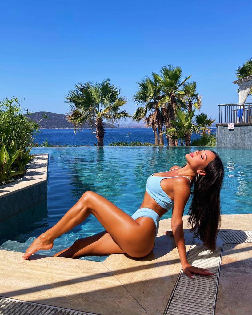 Tanned brunette in a light blue bikini reclining on the edge of an infinity pool with palm trees and the sea behind