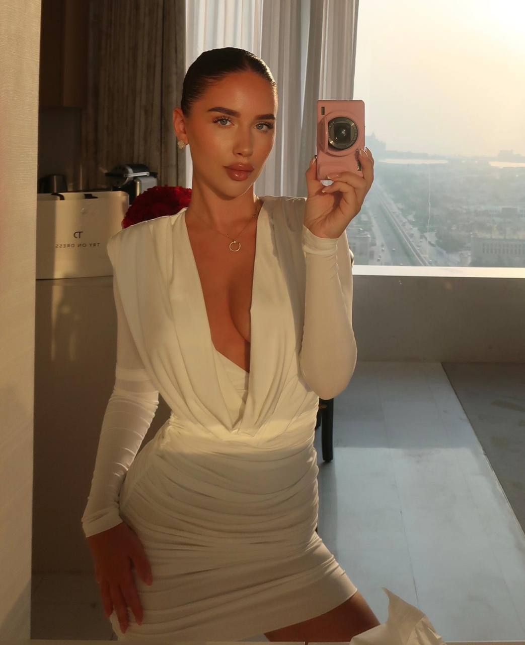 Ultra realistic mirror selfie of a glamorous woman with pale blue eyes, sleek dark hair in a polished ponytail, wearing a fitted white long sleeve bodycon dress with a deep neckline, luxury hotel room, golden hour sunlight through panoramic windows, elegant jewelry, luxury influencer aesthetic, RAW DSLR photo, photorealistic, ultra detailed, 8K.