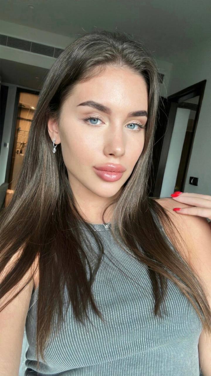 Ultra realistic close-up portrait of a glamorous woman with pale blue eyes, long straight dark brown hair, flawless porcelain skin, naturally full glossy lips, defined eyebrows, wearing a gray sleeveless ribbed top, soft natural window light inside a luxury hotel room, beauty editorial, shallow depth of field, RAW DSLR photo, ultra detailed, photorealistic, 8K.