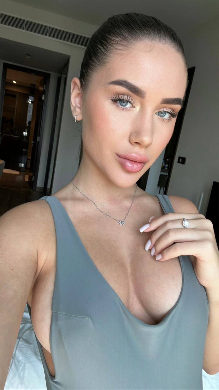 Ultra realistic close-up selfie of a glamorous woman with pale blue eyes, sleek dark hair in a polished ponytail, flawless glowing skin, naturally full glossy lips, wearing a gray deep-neck minimalist top, luxury hotel interior, soft daylight, beauty campaign, Vogue editorial, RAW DSLR photo, ultra detailed, photorealistic, 8K.