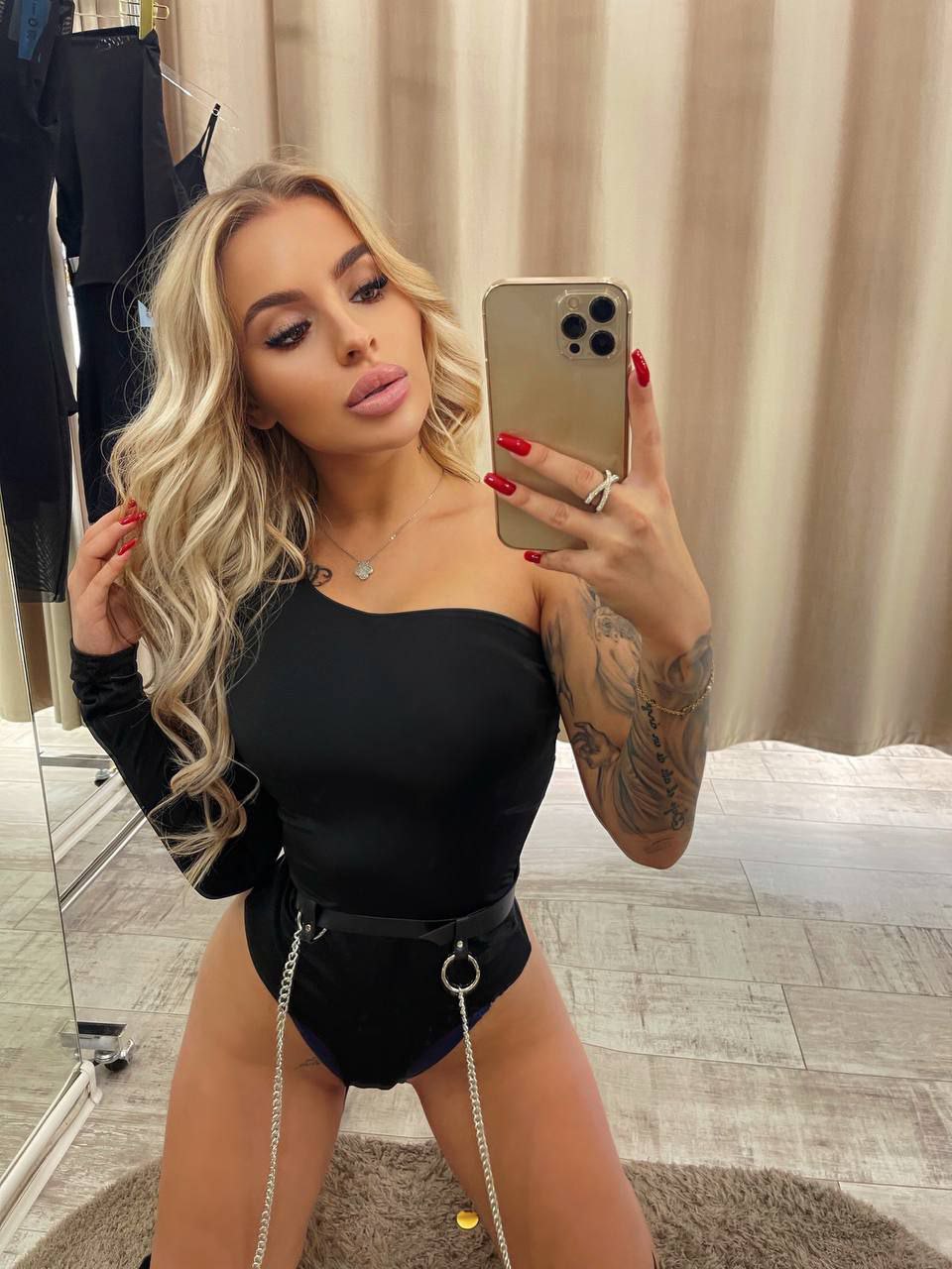 Mirror selfie of a wavy-haired blonde in black one-shoulder bodysuit with chain belt in a fitting room