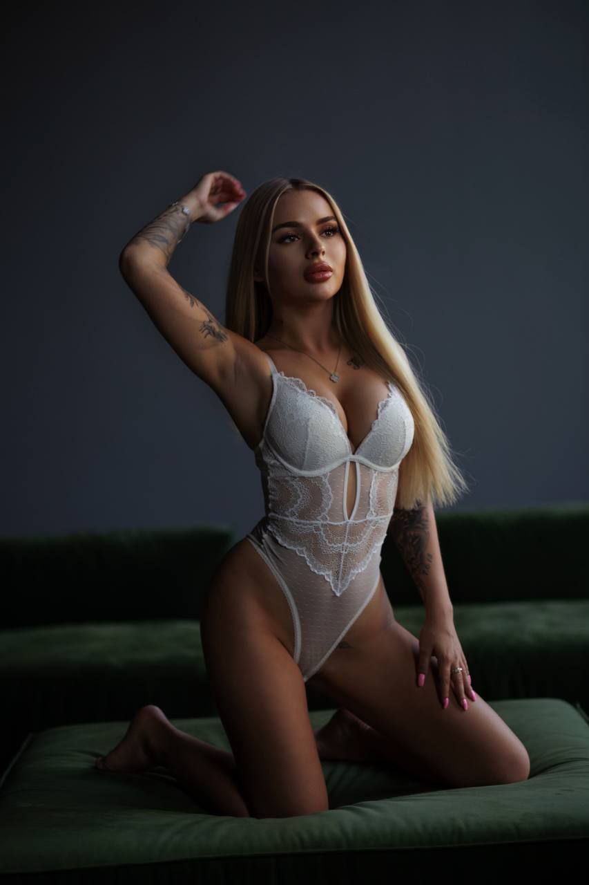 Blonde woman in white lace bodysuit kneeling on a green velvet sofa