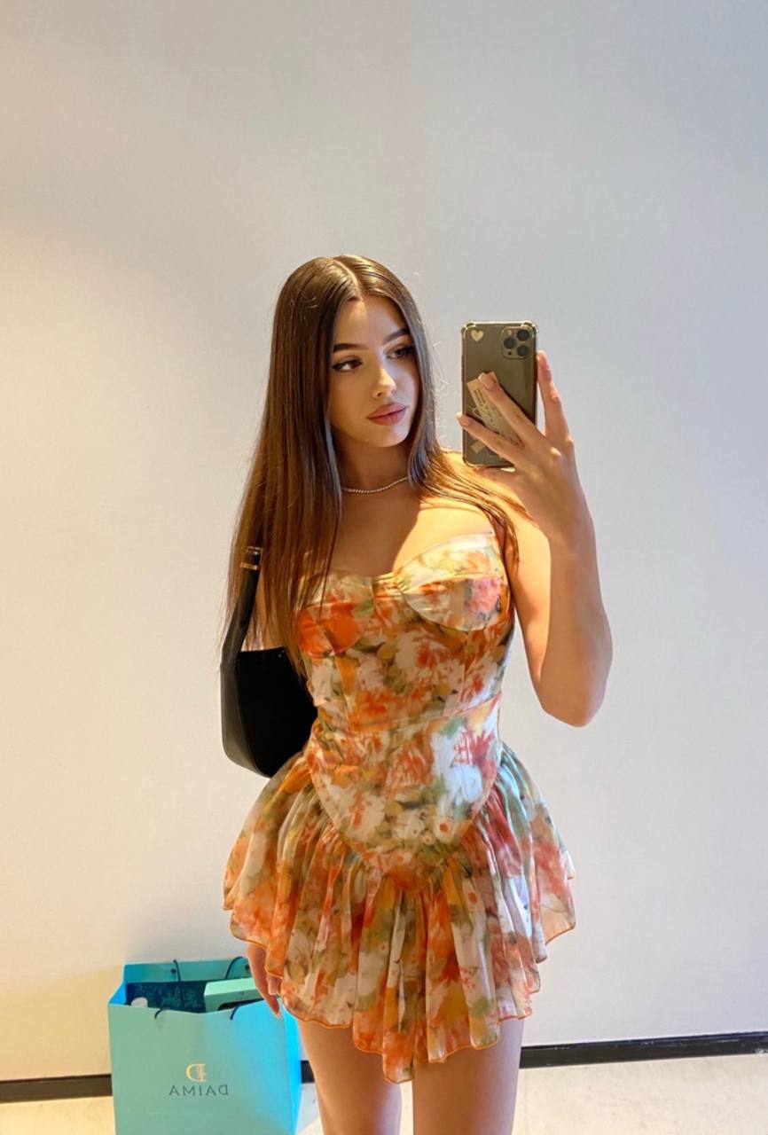 Mirror selfie of a long-haired brunette in a floral corset mini dress with a ruffled skirt and a black shoulder bag