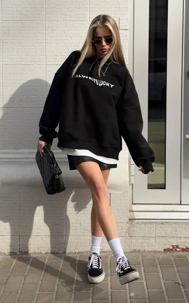 Blonde woman in oversized black sweatshirt, leather mini skirt, white socks and black sneakers posing on the street