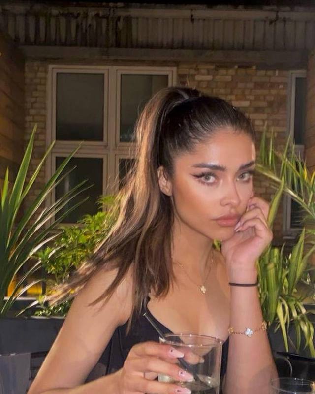 Beautiful brunette fashion model with a sleek high ponytail wearing a black evening top, holding a cocktail in a stylish outdoor café at night with warm ambient lighting