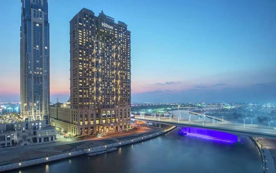 Hilton hotel tower by the Dubai Water Canal at dusk with illuminated purple waterfall bridge
