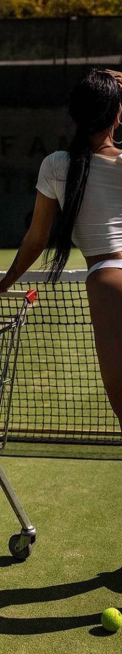 Woman standing on a tennis court beside a cart full of tennis balls and rackets, wearing athletic-inspired casual clothing, photographed from behind in bright outdoor sunlight.