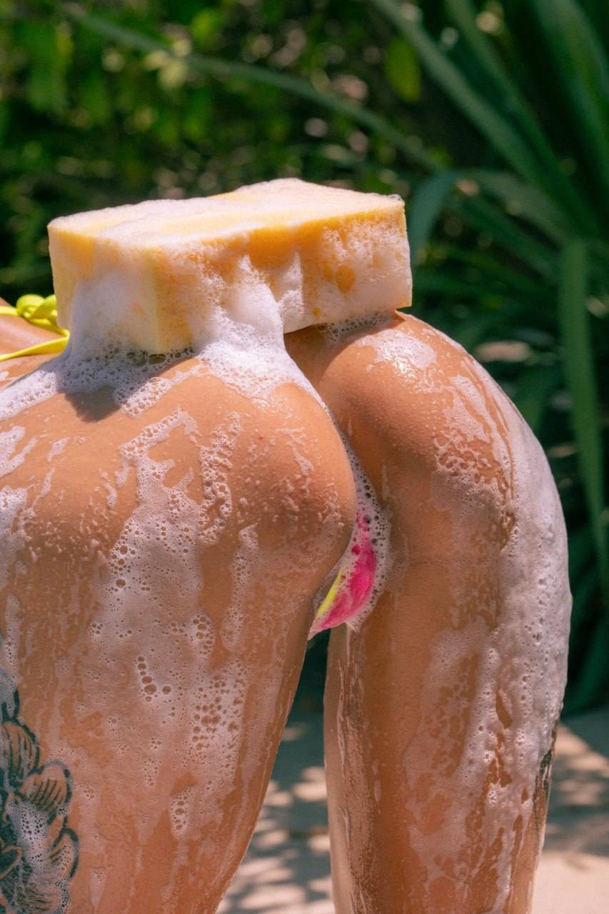 Close-up outdoor lifestyle photo showing a person covered in soap foam with a sponge resting on their lower back, set against lush tropical greenery in bright natural sunlight.