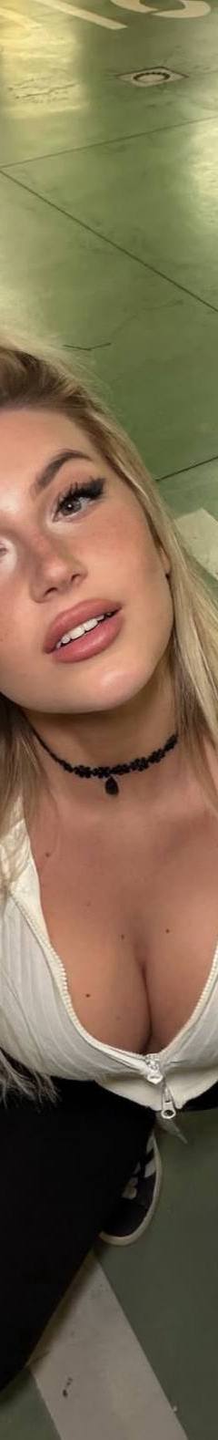 Portrait photo of a blonde woman sitting on the floor of a parking garage, looking up at the camera. She is wearing a white zip-up top, dark pants, sneakers, and a black choker necklace. A hand is lightly touching her hair, and the image is shot from a high angle with painted parking-lot markings visible in the background.