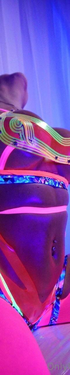 Neon cyberpunk-style portrait of a person wearing fluorescent reflective festival attire under ultraviolet lighting. The image features vivid pink, blue, and purple tones, glowing fabric details, a futuristic aesthetic, and dramatic low-light nightclub or rave-inspired atmosphere.