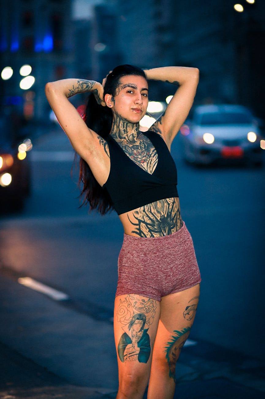 Heavily tattooed woman with dark ponytail in black crop top and pink shorts on a city street at dusk