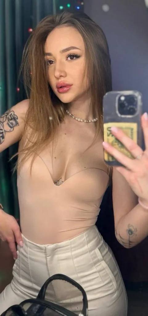 Mirror selfie of a tattooed woman in a beige strapless bodysuit, white trousers and a pearl necklace