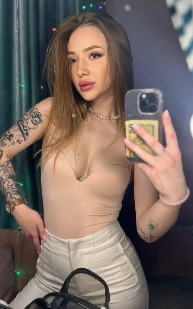 Mirror selfie of a tattooed woman in a beige strapless bodysuit, white trousers and a pearl necklace