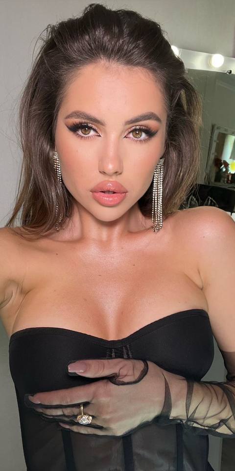Glam selfie of a brunette in a black corset top and sheer mesh gloves with crystal drop earrings at a makeup vanity