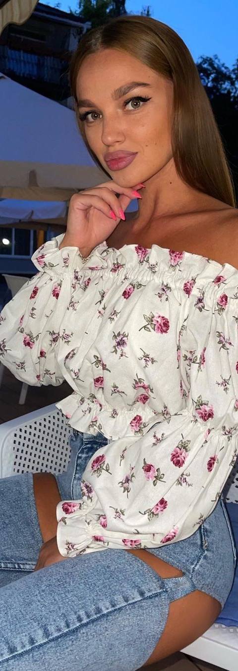 Blonde woman in a white off-shoulder floral blouse and ripped jeans sitting in a chair by an outdoor pool in the evening