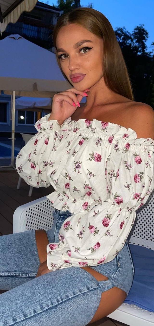 Blonde woman in a white off-shoulder floral blouse and ripped jeans sitting in a chair by an outdoor pool in the evening