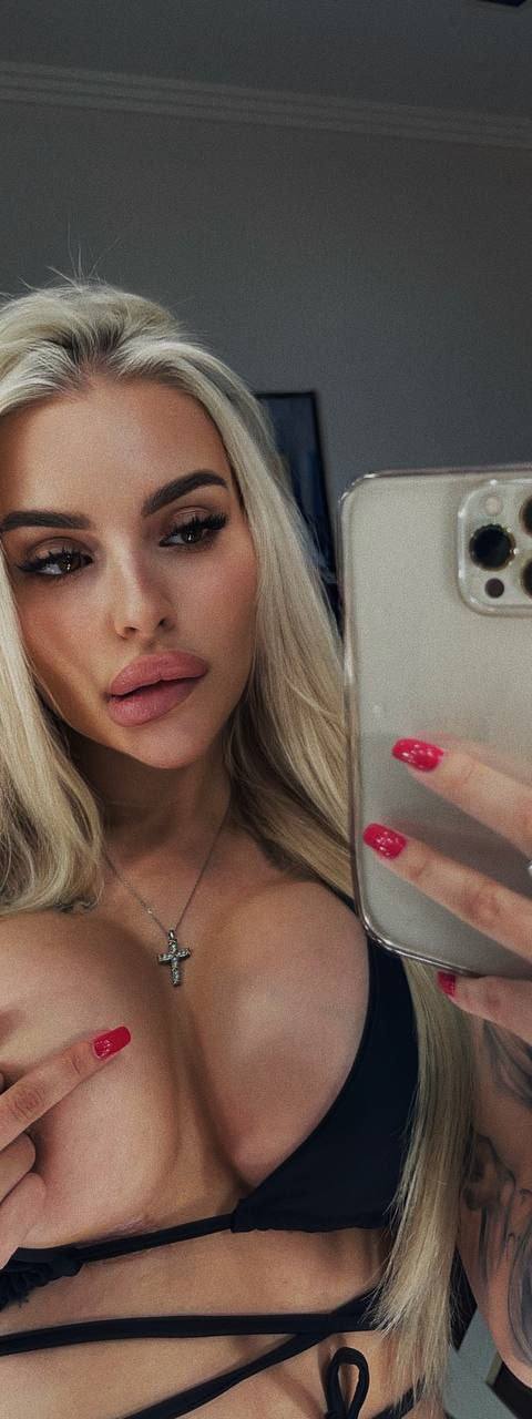 Mirror selfie of a long-haired blonde woman in black wrap top holding a phone
